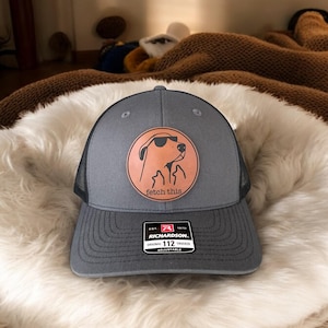 Puede incluir: Gorra de camionero gris con la parte trasera de malla negra y un parche de cuero con un perro con gafas de sol. El parche tiene las palabras "fetch this". La gorra es ajustable.