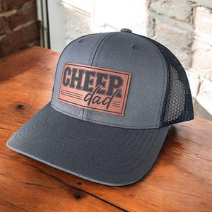 May include: A grey and black trucker hat with a brown leather patch that says "CHEER dad" in black lettering.