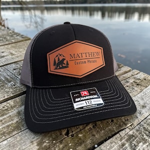 May include: Black and grey trucker hat with a brown leather patch. The patch features the name "MATTHEW", a bear, trees, and the words "Custom Phrase". The hat has a Richardson 112 tag.
