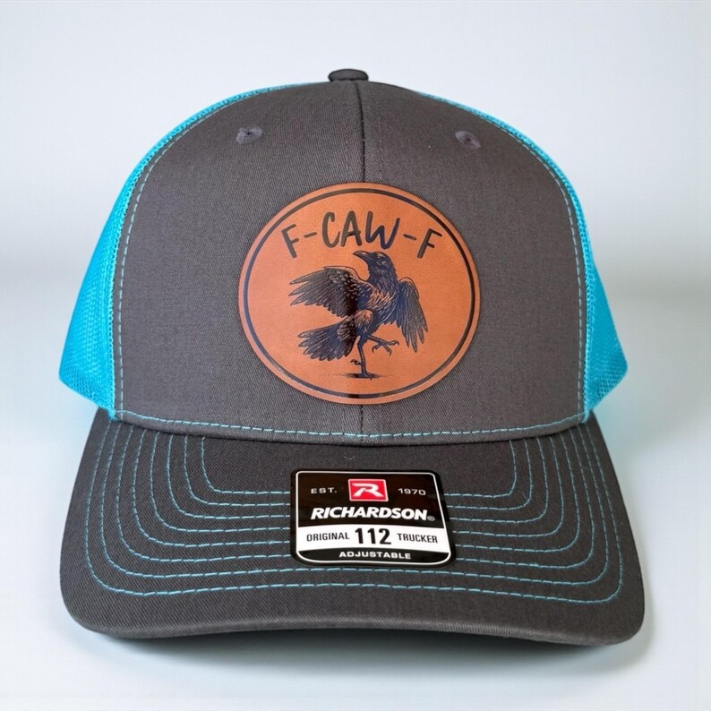 May include: Gray and blue trucker hat with a leather patch featuring a crow illustration and the text "F-CAW-F." The hat has a curved brim and adjustable closure. The Richardson brand label is visible.