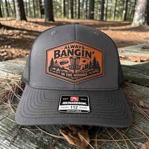 May include: A gray trucker hat with a black mesh back and a curved brim. The front features a brown patch with the text "ALWAYS BANGIN' IN THE WOODS" and a disc golf basket graphic. The hat has an adjustable strap.