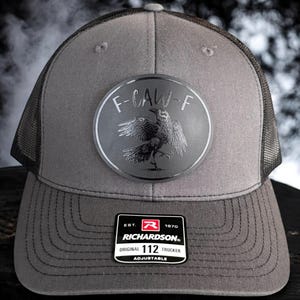 May include: Gray trucker hat with a mesh back and a curved brim. The hat features a circular patch with a raven design and the text "F-CAW-F". A rectangular tag reads "RICHARDSON ORIGINAL 112 TRUCKER ADJUSTABLE".