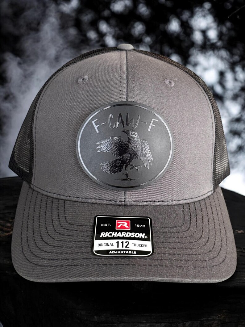 May include: Gray trucker hat with a mesh back and a curved brim. The hat features a circular patch with a raven design and the text "F-CAW-F". A rectangular tag reads "RICHARDSON ORIGINAL 112 TRUCKER ADJUSTABLE".