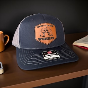Può includere: Cappellino da camionista blu navy con retro in rete bianca e visiera curva. Il cappello presenta una toppa in pelle con due illustrazioni di wombat e la scritta "HAND TO HAND WOMBAT". Il cappellino Richardson 112 è regolabile.