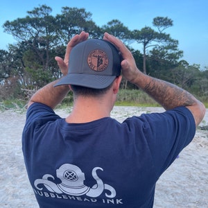 May include: A person wearing a dark gray t-shirt with a white octopus and diving helmet graphic and the text "Bubblehead Ink". The person is also wearing a dark gray and black trucker hat with a brown leather patch that says "Support Your Local Pole Dancer".