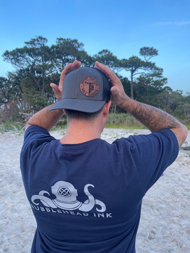 May include: A person wearing a dark gray t-shirt with a white octopus and diving helmet graphic and the text "Bubblehead Ink". The person is also wearing a dark gray and black trucker hat with a brown leather patch that says "Support Your Local Pole Dancer".