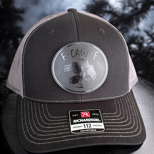 May include: A black and gray Richardson 112 trucker hat with a mesh back. The hat features a circular patch with the text "F-CAW-F" and a raven illustration. The hat is adjustable.