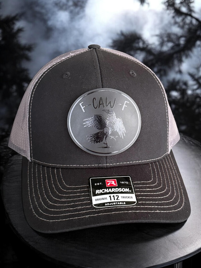 May include: A black and gray Richardson 112 trucker hat with a mesh back. The hat features a circular patch with the text "F-CAW-F" and a raven illustration. The hat is adjustable.