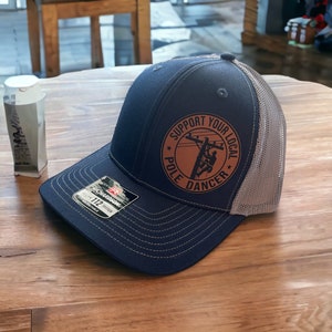 May include: A navy blue and gray trucker hat with a brown leather patch that says "Support Your Local Pole Dancer".
