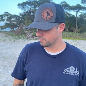 May include: A gray and black trucker hat with a brown leather patch that says "Support Your Local Pole Dancer". The hat is worn by a person wearing a navy blue t-shirt with a white octopus and diving helmet graphic that says "Bubblehead Ink".