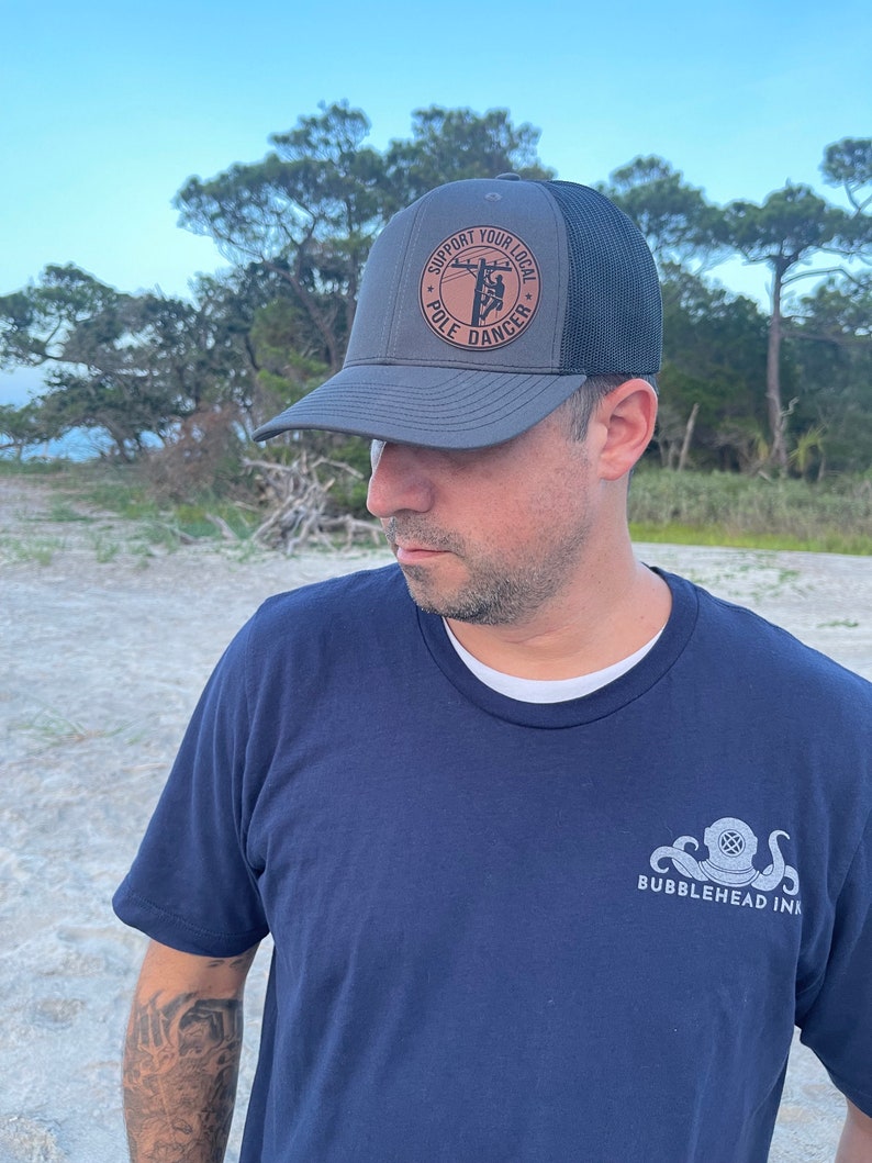 May include: A gray and black trucker hat with a brown leather patch that says "Support Your Local Pole Dancer". The hat is worn by a person wearing a navy blue t-shirt with a white octopus and diving helmet graphic that says "Bubblehead Ink".