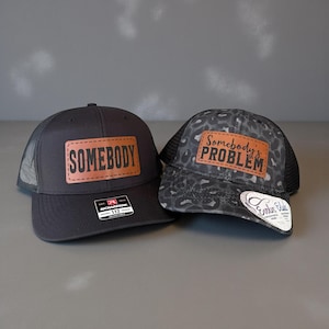 Puede incluir: Dos gorras de camionero. Una es negra con un parche de cuero marrón que dice "SOMEBODY". La otra es de estampado de leopardo gris con un parche de cuero marrón que dice "Somebody's PROBLEM". Ambas tienen la parte trasera de malla.