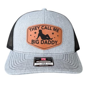 Op de afbeelding: Grijze truckerpet met een zwarte mesh achterkant. De voorkant heeft een bruine leren patch met de tekst "THEY CALL ME BIG DADDY" en een silhouet. De pet heeft een gebogen klep en een verstelbare band.