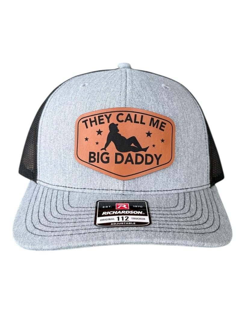 Op de afbeelding: Grijze truckerpet met een zwarte mesh achterkant. De voorkant heeft een bruine leren patch met de tekst "THEY CALL ME BIG DADDY" en een silhouet. De pet heeft een gebogen klep en een verstelbare band.