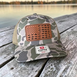 May include: A camouflage trucker hat featuring a rectangular leather patch with a flag design composed of small fish silhouettes. The hat has a mesh back and a sticker on the brim that reads R 112PFP PRINTED S PANEL OSFM.