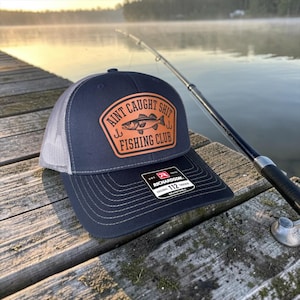 May include: A navy blue and gray trucker hat with the text "Aint Caught Shit Fishing Club" in a brown patch. The hat has a curved brim and an adjustable strap. A fishing rod is in the background.