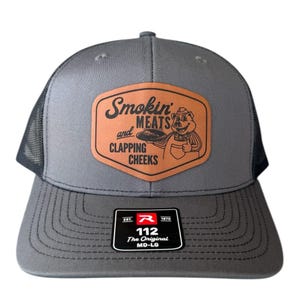 May include: Gray trucker hat with a mesh back and a curved brim. The front features a brown leather patch with the text "Smokin' MEATS and CLAPPING CHEEKS" and a cartoon pig. The hat has a black tag that says "112 The Original MD-LG".