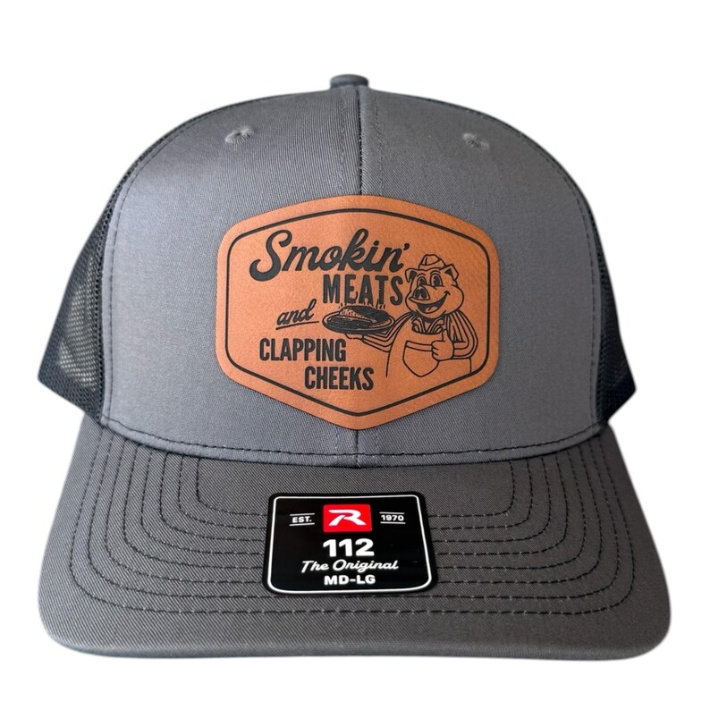 May include: Gray trucker hat with a mesh back and a curved brim. The front features a brown leather patch with the text "Smokin' MEATS and CLAPPING CHEEKS" and a cartoon pig. The hat has a black tag that says "112 The Original MD-LG".
