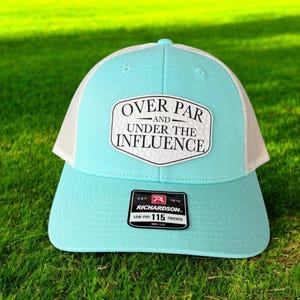 May include: A light blue and white trucker hat with a white patch that says "Over Par and Under the Influence". The hat is made by Richardson and has a size tag that says "Low-Pro 115 Trucker".