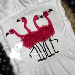 May include: Embroidered design of three pink flamingos with black legs and beaks on a white shirt. The flamingos are standing on top of each other, with the top flamingo's head facing forward and the other two flamingos' heads facing sideways. The shirt has a ruffled collar and a small logo that says "The Fancy Flamingo Boutique".