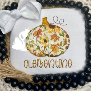 May include: White onesie with an embroidered autumn-themed pumpkin with fall leaves and sunflowers. The onesie also has the text "Clementine" embroidered on it.