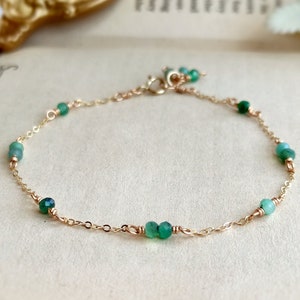 May include: A delicate gold chain bracelet with small green gemstone beads. The bracelet is on a light brown surface.