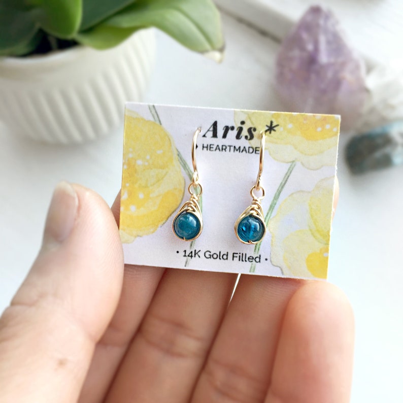 May include: Gold-filled earrings featuring teal gemstones in a teardrop shape. The earrings are presented on a card with the text "Aris Heartmade" and "14K Gold Filled."