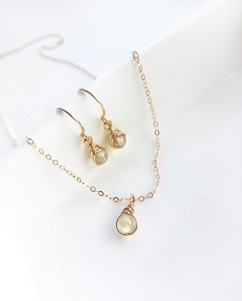 May include: A gold-colored jewelry set featuring a necklace and earrings. The necklace has a small, teardrop-shaped pendant. The earrings and pendant have a wire-wrapped design, with a small, round, light-colored stone in each.