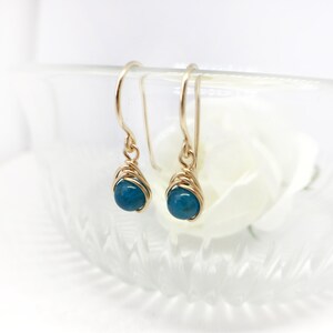 May include: A pair of gold-coloured wire-wrapped earrings. Each earring features a small, round, teal bead. The earrings have a simple hook design and are set against a soft, blurred background.