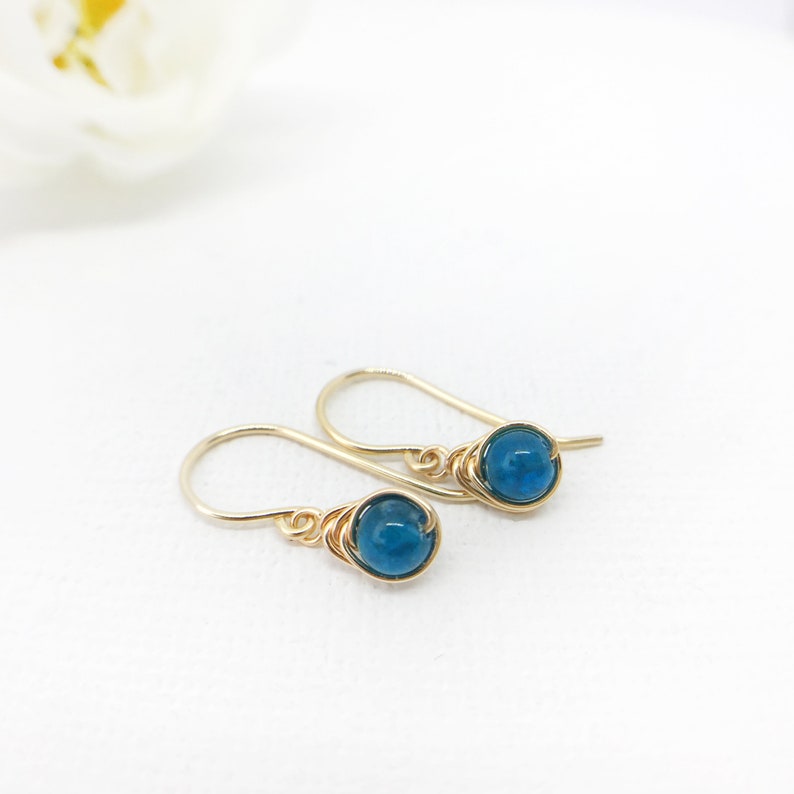 May include: A pair of gold-coloured earrings featuring blue gemstone beads. The beads are wire-wrapped and hang from curved ear wires. The earrings are set against a white backdrop.