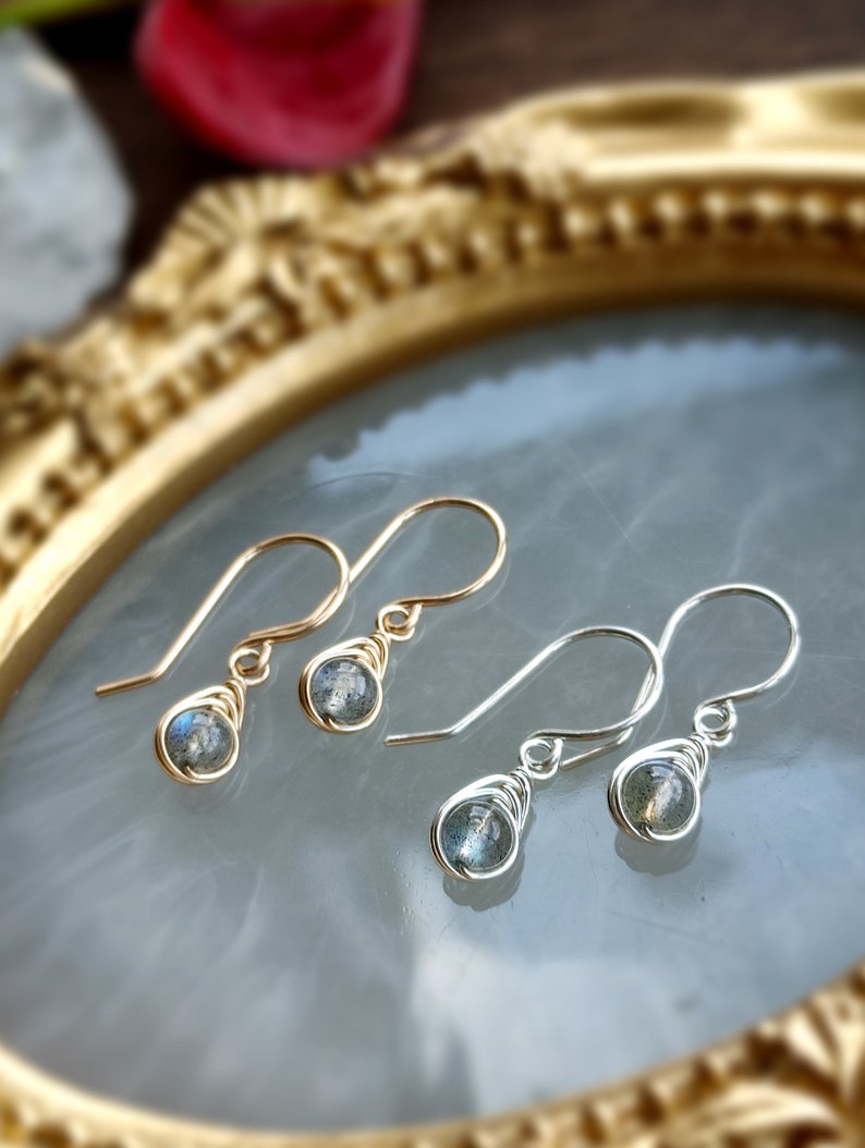May include: Two pairs of dangle earrings. One pair features gold-colored wire and the other silver-colored wire. Each earring has a small, round, gray gemstone wrapped in wire. The earrings are displayed on a light gray surface.