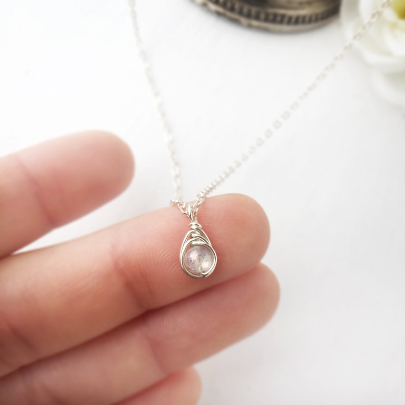 May include: A delicate silver necklace featuring a small, teardrop-shaped pendant. The pendant is a wire-wrapped, iridescent gemstone, likely a moonstone or labradorite. The necklace is held by a person's fingers, showcasing the jewelry's size and design.