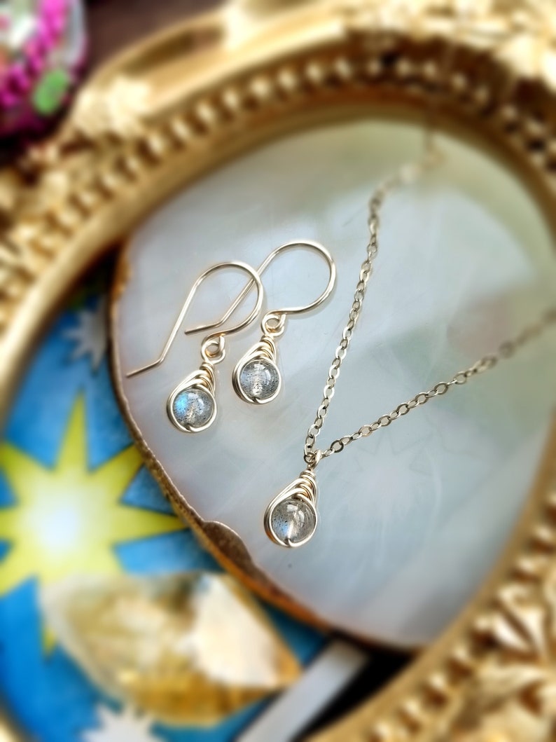 May include: A gold-colored jewelry set featuring a necklace and matching earrings. Each piece has a small, round, iridescent stone wrapped in gold wire. The earrings are drop style, and the necklace has a delicate chain.