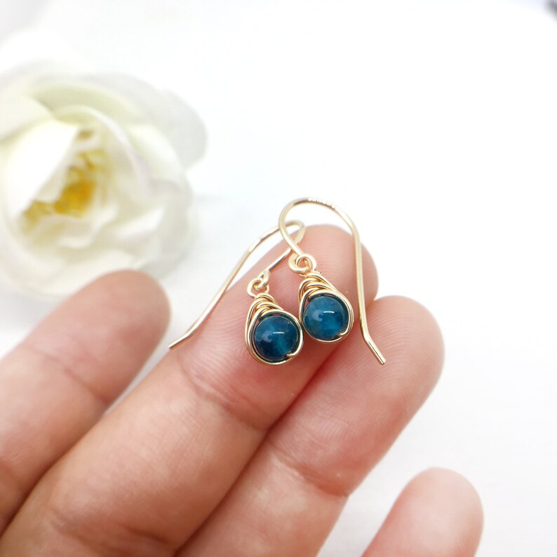 May include: Gold-coloured wire-wrapped earrings featuring deep teal, teardrop-shaped beads. The earrings are set against a soft white backdrop, with a blurred white flower in the upper left corner. The earrings are a delicate piece of jewellery.