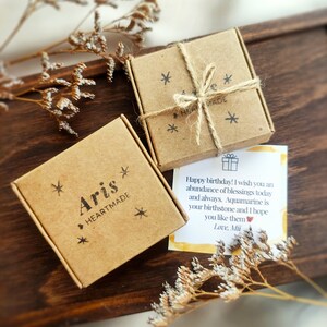 May include: Two small, brown cardboard boxes tied with twine, one labeled "Aris HEARTMADE" and the other with asterisks. A handwritten birthday card with text is visible. The boxes and card rest on a dark wooden surface, with dried flowers.