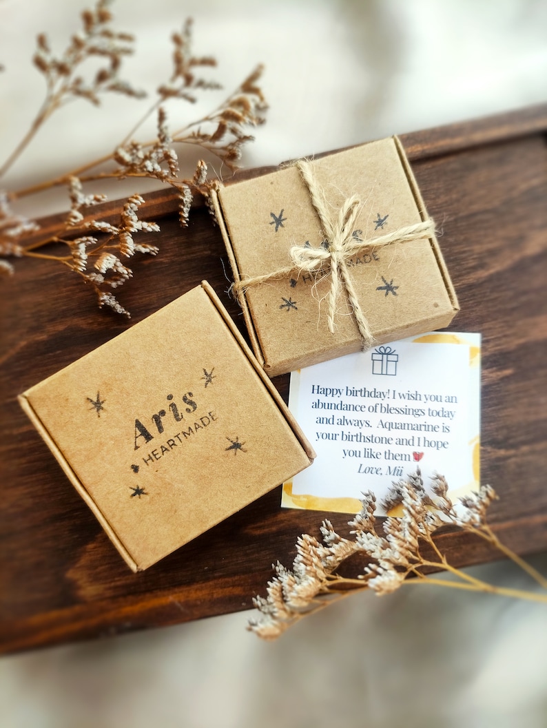 May include: Two small, brown cardboard boxes tied with twine, one labeled "Aris HEARTMADE" and the other with asterisks. A handwritten birthday card with text is visible. The boxes and card rest on a dark wooden surface, with dried flowers.