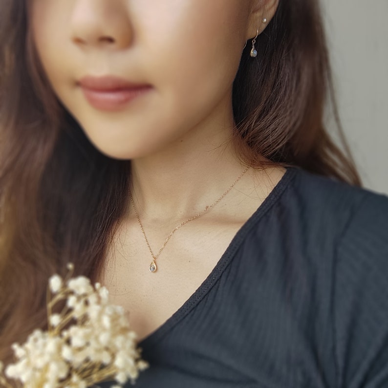 May include: A gold necklace with a small, teardrop-shaped pendant and matching earrings. The jewelry is delicate and elegant, with a subtle sparkle. The necklace is worn on a person wearing a black top.