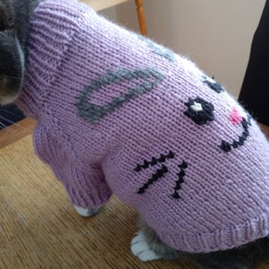 Puede incluir: Un suéter de punto lavanda para un gato con un diseño de cara de conejo blanco y negro. El suéter tiene orejas grises y una nariz rosa.