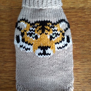 Könnte beinhalten: Ein beiger Strickpullover für Hunde mit einem Tigergesichtsdesign in Gelb, Schwarz und Weiß.
