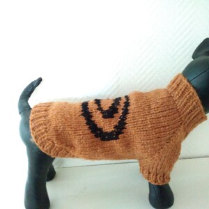 Puede incluir: Un suéter de punto marrón para perro con un diseño geométrico negro. El suéter está en un maniquí de perro negro.