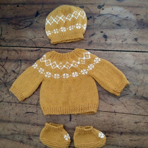 May include: A mustard yellow knitted baby outfit with a white geometric pattern. The outfit includes a hat, sweater, and booties.