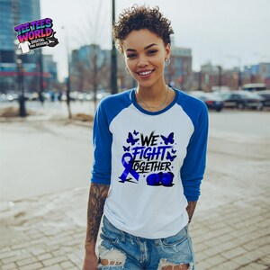May include: A white and blue baseball tee with the text "WE FIGHT TOGETHER" in blue, along with a blue ribbon and butterflies. The shirt has blue sleeves and is worn by a person in blue denim shorts.