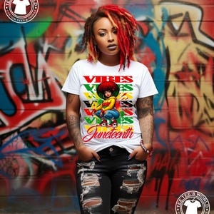 May include: White t-shirt with the word "Vibes" in red, green, and yellow, and a graphic of a woman. The word "Juneteenth" is written in cursive. The background is a graffiti wall.