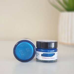 May include: Two small glass jars of blue pigment, one upright and one on its side. The upright jar has a black lid and a label that reads "Offshore Wave". The pigment inside the jars is a vibrant, shimmering blue.