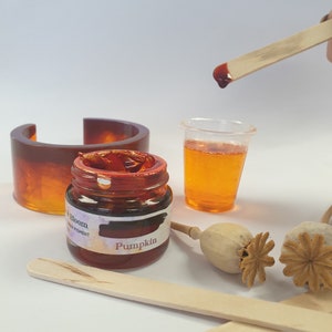 May include: A small glass jar of orange resin pigment labeled "Bloom Resin Pigment Pumpkin". A wooden craft stick is dipping into the jar, and a clear plastic cup filled with orange resin sits next to it. Two dried poppy seed pods are on the table.