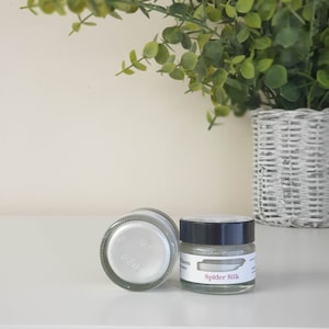 May include: Two small jars of white shimmery paint. The jar on the right has a black lid and the text "Spider Silk" printed on the label.