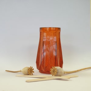 May include: A clear orange glass vase with a textured surface. The vase is sitting on a white surface with two dried poppy seed pods.
