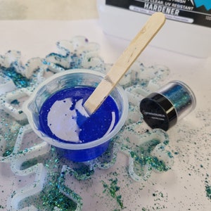 May include: A clear plastic cup with a blue resin mixture and a wooden stirring stick. A small jar of iridescent green glitter is next to the cup. The resin and glitter are on a white surface with white plastic snowflake shapes.