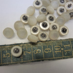 May include: A collection of 18 white, round, iridescent buttons with silver metal shanks. The buttons are arranged on a white surface with a green measuring tape.