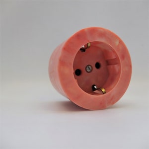 Peut inclure: Une prise électrique en plastique rose avec un motif marbré. La prise a trois trous ronds et des contacts métalliques dorés.